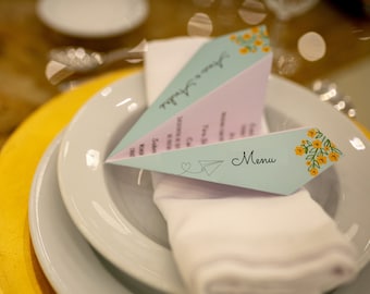 May include: A light blue and pink paper airplane menu with yellow flowers and the word "Menu" printed on it. The menu is placed on a white napkin on a white plate.