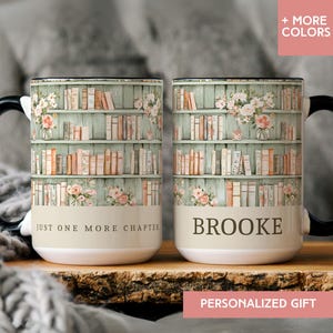 May include: Two white ceramic mugs with black handles. The mugs feature a bookshelf design with books and floral accents. One mug says "JUST ONE MORE CHAPTER", the other says "BROOKE". The mugs are on a wooden surface.