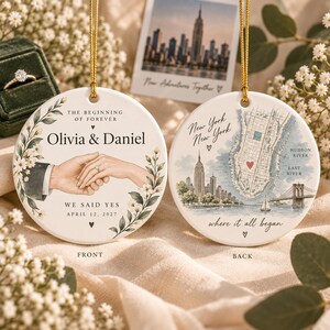 May include: Two round white ceramic ornaments with gold hanging strings. The front features an illustration of two hands holding, the names Olivia & Daniel, and the text The Beginning of Forever and We Said Yes April 12, 2027. The back shows a map of New York.