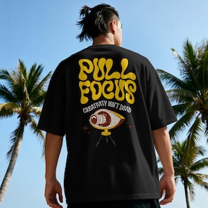 Könnte beinhalten: Schwarzes T-Shirt mit dem gelben Schriftzug "PULL FOCUS", dazu "CREATIVITY ISN'T DEAD" und eine augenförmige Kamera-Grafik. Das T-Shirt wird von einer Person im Freien getragen, mit Palmen im Hintergrund.
