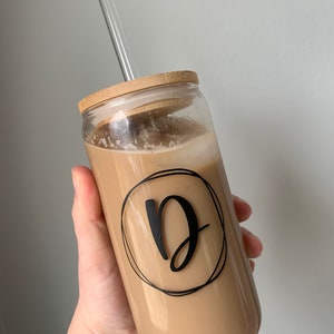 Op de afbeelding: Een heldere glazen beker gevuld met een romige drank, met een bamboe deksel en een helder glazen rietje. De beker heeft een zwarte letter "D" in een cirkelvormig ontwerp. De drank wordt in een hand gehouden.
