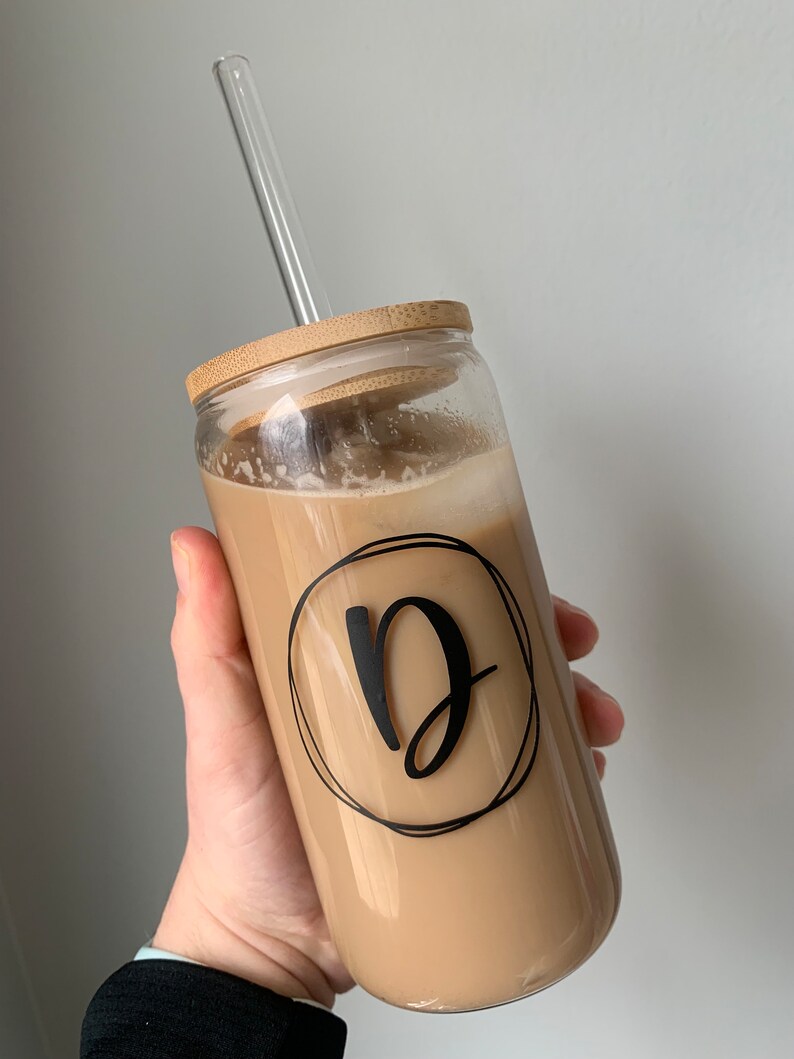Op de afbeelding: Een heldere glazen beker gevuld met een romige drank, met een bamboe deksel en een helder glazen rietje. De beker heeft een zwarte letter "D" in een cirkelvormig ontwerp. De drank wordt in een hand gehouden.