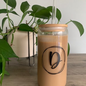 Può includere: Un bicchiere trasparente pieno di una bevanda marrone chiaro, con un coperchio di bambù e una cannuccia di vetro trasparente. Il bicchiere ha un design circolare nero con la lettera "D". Una pianta verde e frondosa è sullo sfondo.