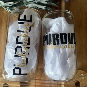 May include: Two clear glass can-shaped cups with white tissue paper inside. The cups have the words "PURDUE" and "boilermakers" in black and gold lettering. The cups are on a wooden surface.