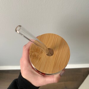 Op de afbeelding: Een glazen beker met een bamboe deksel en een doorzichtige glazen rietje. De deksel heeft een rond gat voor het rietje. De beker wordt in een hand gehouden, met een zwarte mouw zichtbaar.