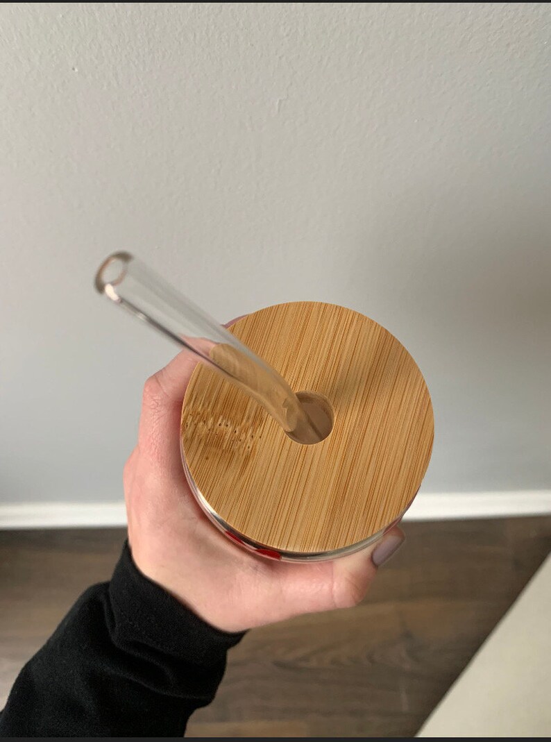 Op de afbeelding: Een glazen beker met een bamboe deksel en een doorzichtige glazen rietje. De deksel heeft een rond gat voor het rietje. De beker wordt in een hand gehouden, met een zwarte mouw zichtbaar.
