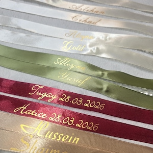 Pu&ograve; includere: Primo piano di diversi nastri di raso in vari colori, tra cui bianco, verde, bordeaux e beige. Ogni nastro &egrave; inciso con lettere dorate, con nomi e date. I nastri sono disposti vicini, creando un effetto a strati.