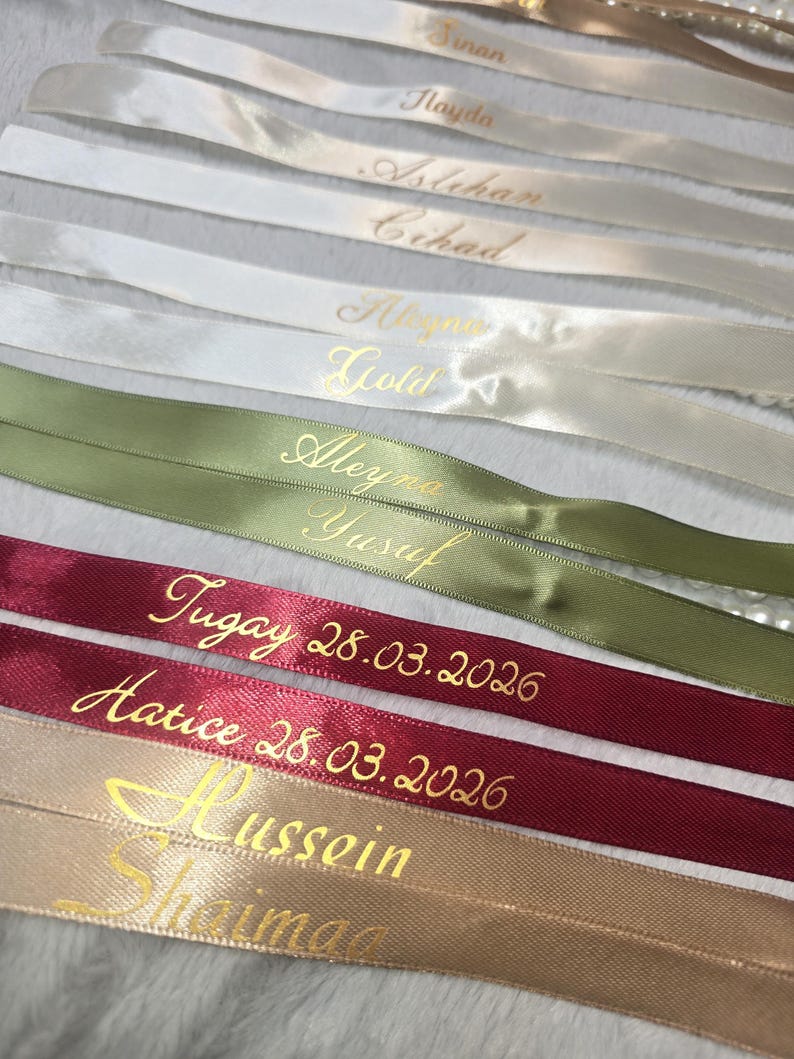 Pu&ograve; includere: Primo piano di diversi nastri di raso in vari colori, tra cui bianco, verde, bordeaux e beige. Ogni nastro &egrave; inciso con lettere dorate, con nomi e date. I nastri sono disposti vicini, creando un effetto a strati.