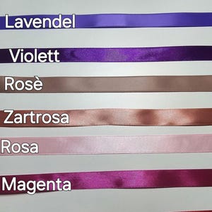 Pu&ograve; includere: Una collezione di nastri di raso in vari colori, tra cui lavanda, viola, rosa e magenta. Ogni nastro &egrave; etichettato con il suo nome di colore in tedesco. I nastri sono disposti orizzontalmente su uno sfondo chiaro.