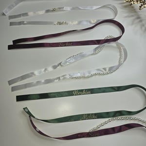 Pu&ograve; includere: Assortimento di nastri di raso personalizzati in bianco, bordeaux e verde, ciascuno con un nome in lettere dorate. Alcuni nastri sono adornati con una fila di perle. I nastri sono disposti su una superficie bianca.