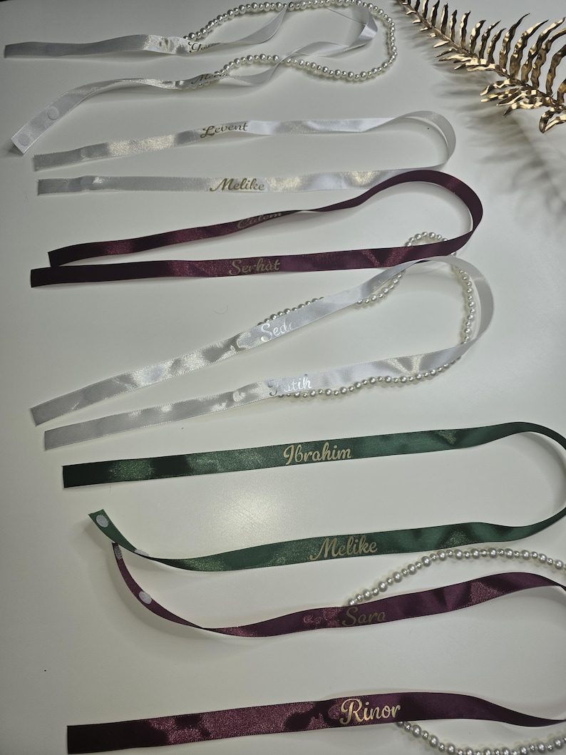 Pu&ograve; includere: Assortimento di nastri di raso personalizzati in bianco, bordeaux e verde, ciascuno con un nome in lettere dorate. Alcuni nastri sono adornati con una fila di perle. I nastri sono disposti su una superficie bianca.