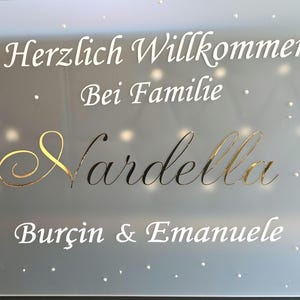 Può includere: Un'insegna in vetro smerigliato con le parole "Herzlich Willkommen Bei Familie" in bianco e "Nardella" in corsivo dorato. Anche i nomi "Burçin & Emanuele" sono in bianco. Piccoli punti bianchi sono sparsi sulla superficie.