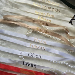 Pu&ograve; includere: Assortimento di nastri di raso personalizzati in vari colori, tra cui verde, bianco, beige e rosso. Ogni nastro presenta scritte dorate con nomi e date. Alcuni nastri sono adornati con perle.