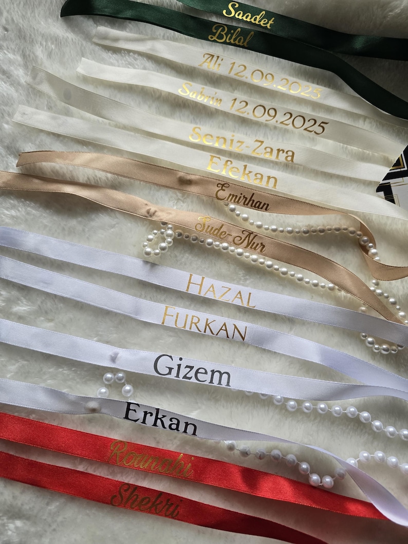 Pu&ograve; includere: Assortimento di nastri di raso personalizzati in vari colori, tra cui verde, bianco, beige e rosso. Ogni nastro presenta scritte dorate con nomi e date. Alcuni nastri sono adornati con perle.