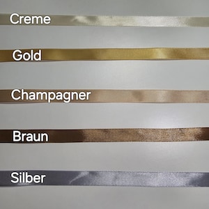 Pu&ograve; includere: Una selezione di nastri di raso in vari colori, tra cui bianco, crema, oro, champagne, marrone, argento e nero. Ogni nastro &egrave; etichettato con il suo nome di colore in tedesco. I nastri sono disposti orizzontalmente.