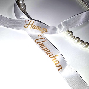 Pu&ograve; includere: Nastro di raso bianco con scritte dorate che recitano "Hamza" e "Ummihan". &Egrave; visibile anche un filo di perle bianche. Il nastro viene probabilmente utilizzato per confezioni regalo o occasioni speciali.