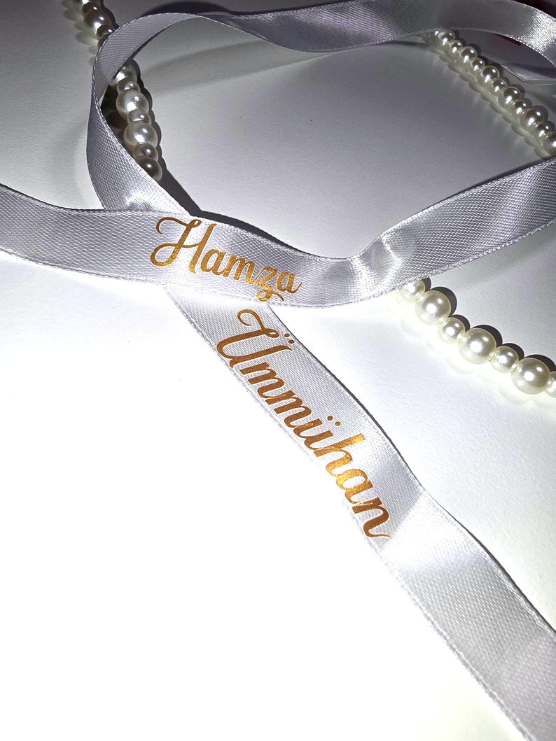 Pu&ograve; includere: Nastro di raso bianco con scritte dorate che recitano "Hamza" e "Ummihan". &Egrave; visibile anche un filo di perle bianche. Il nastro viene probabilmente utilizzato per confezioni regalo o occasioni speciali.