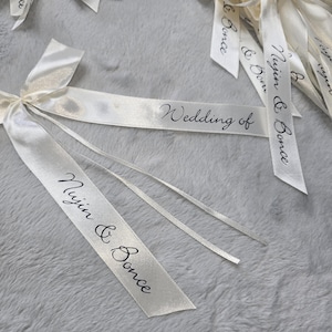 Puede incluir: Cintas de raso color marfil con texto de caligrafía negra. Una cinta dice "Wedding of", mientras que otras muestran "Nujin & Bonce". Varias cintas están atadas en lazos, lo que sugiere su uso como recuerdos de boda o decoraciones.