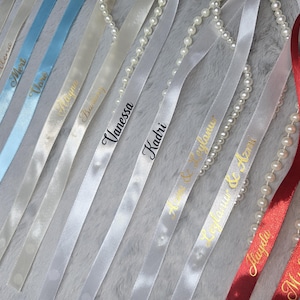 Pu&ograve; includere: Assortimento di nastri di raso in azzurro, bianco e rosso, personalizzati con nomi in corsivo dorato e nero. Alcuni nastri sono adornati con fili di perle, creando un accessorio elegante e personalizzato.