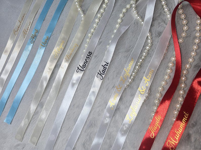 Pu&ograve; includere: Assortimento di nastri di raso in azzurro, bianco e rosso, personalizzati con nomi in corsivo dorato e nero. Alcuni nastri sono adornati con fili di perle, creando un accessorio elegante e personalizzato.