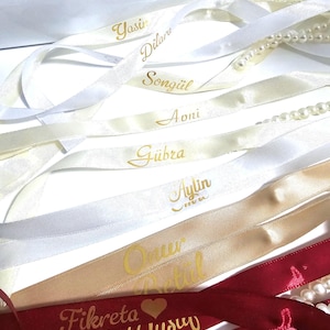Pu&ograve; includere: Un assortimento di nastri di raso bianchi, crema, oro e bordeaux, ciascuno con un nome in oro. Alcuni nastri sono abbinati a fili di perle. I nastri sono disposti su una superficie bianca.
