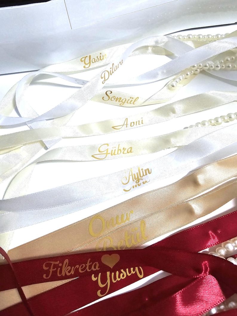 Pu&ograve; includere: Un assortimento di nastri di raso bianchi, crema, oro e bordeaux, ciascuno con un nome in oro. Alcuni nastri sono abbinati a fili di perle. I nastri sono disposti su una superficie bianca.