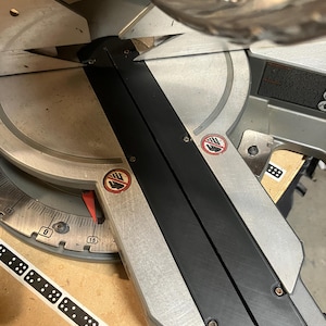 May include: A close-up of a miter saw, a power tool used for making precise angle cuts. The saw features a silver blade guard, a black base, and a measurement scale. The blade is partially visible, and the saw is set on a wooden surface.