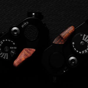 May include: Close-up of a black camera with wooden accents. The camera features dials with numbers and settings, including ASA/ISO and aperture controls. The wooden accents provide a contrast to the black body.