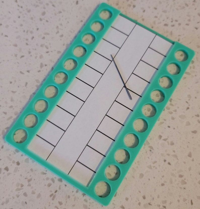 May include: A turquoise plastic thread organizer with a gridded white card and a single needle. The organizer has 18 round holes on each side.