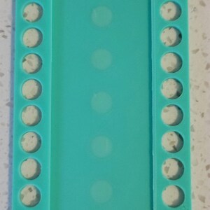 May include: A turquoise plastic bead organizer with 10 rows of circular holes on each side. The organizer is designed to hold beads, charms, or other small crafting supplies.