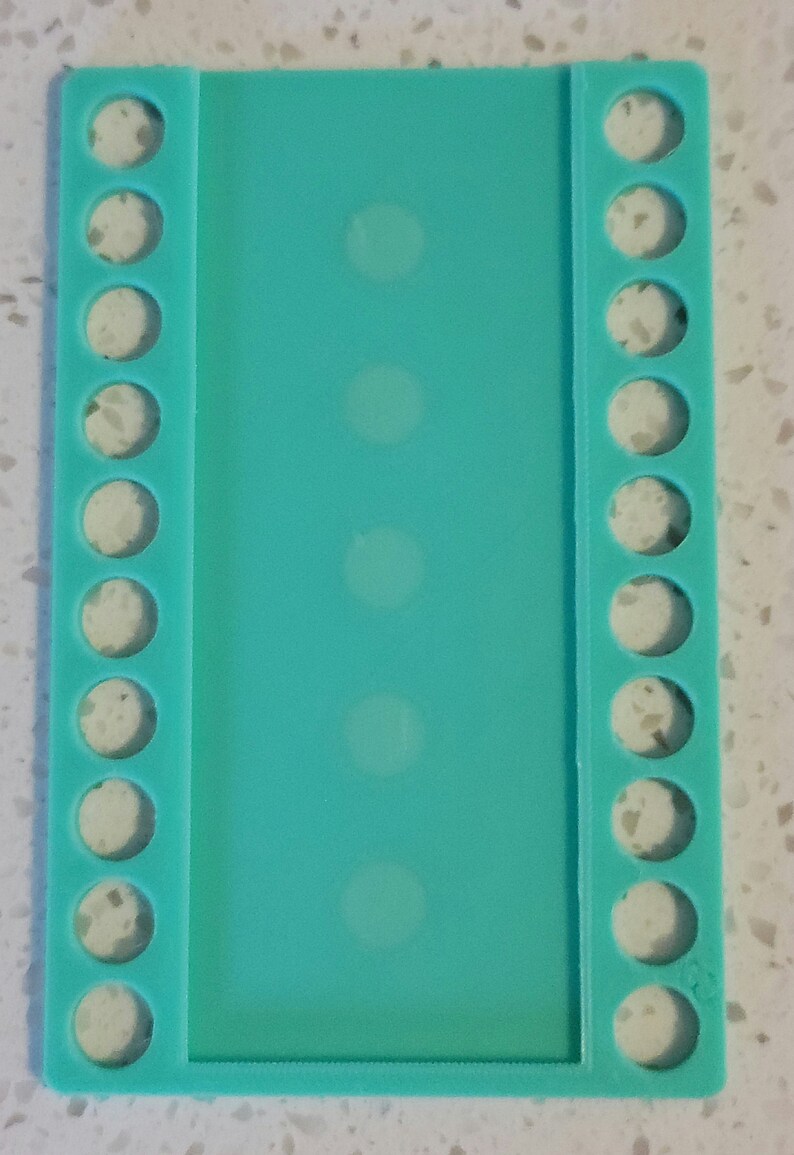 May include: A turquoise plastic bead organizer with 10 rows of circular holes on each side. The organizer is designed to hold beads, charms, or other small crafting supplies.
