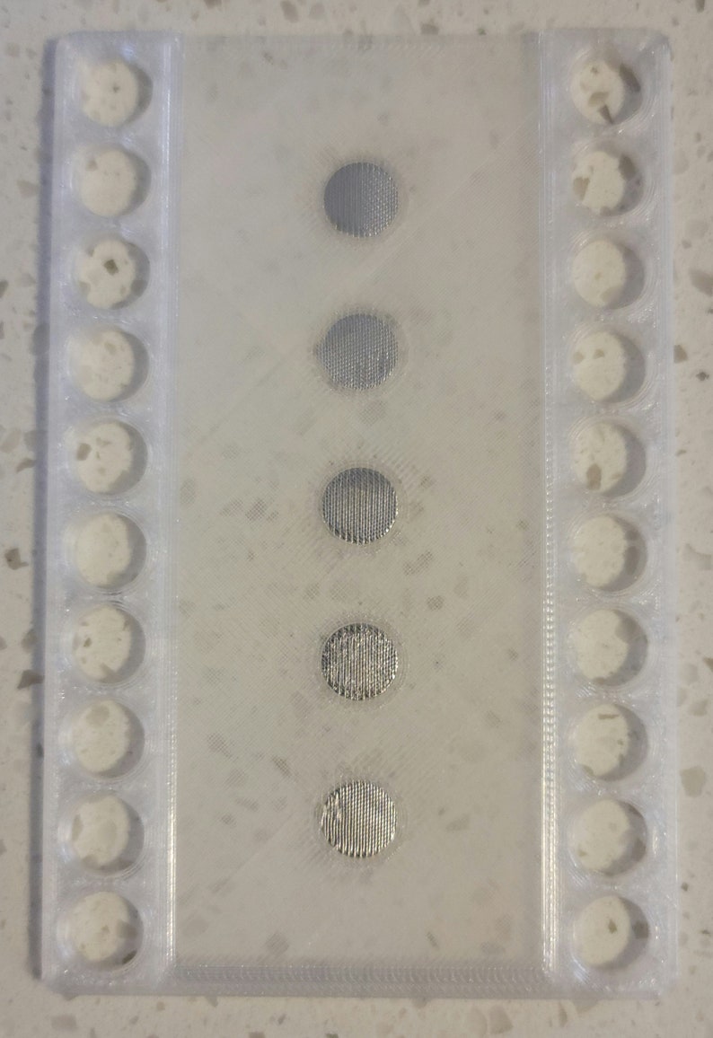 May include: A white plastic tray with 10 round holes and 5 silver metal discs with a textured surface.