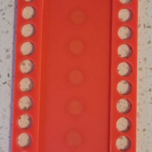 May include: A bright orange plastic bead organizer with rows of circular holes. The organizer has a total of 15 holes.