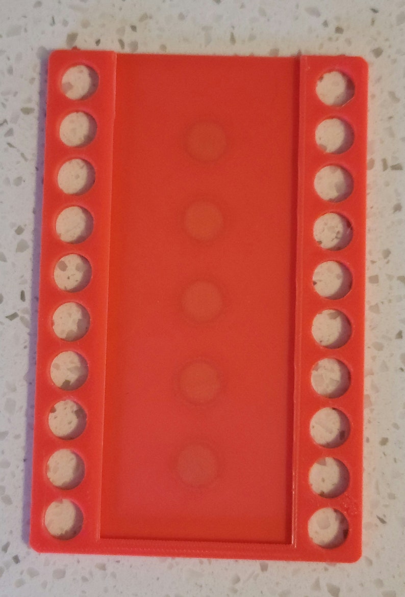 May include: A bright orange plastic bead organizer with rows of circular holes. The organizer has a total of 15 holes.