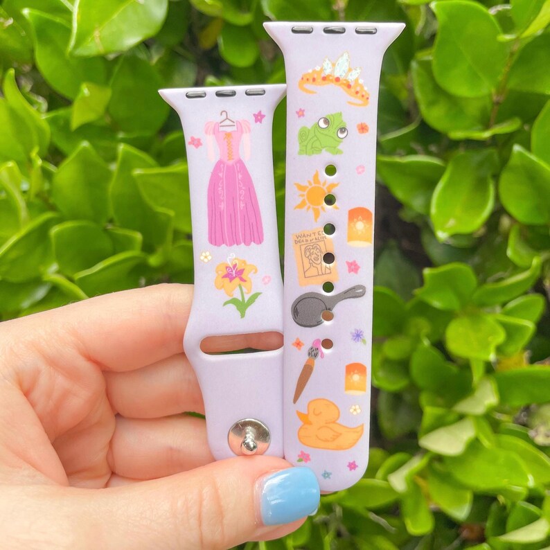 May include: Two light purple watch bands with colorful graphics. One band features a pink dress, a yellow flower, and stars. The other band has a frog, a sun, a frying pan, a duck, and a wanted poster.