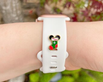 Puede incluir: Una correa blanca para smartwatch con un dije de calcetín navideño dorado y esmaltado. El dije presenta un diseño de calcetín rojo, verde y blanco con una cabeza de Mickey Mouse y las palabras "Merry Christmas".