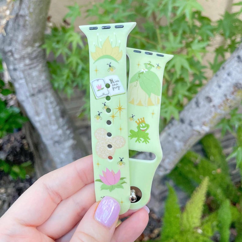 May include: Green Apple Watch band with a light green background and a white design featuring a frog, a princess, a book, and a flower. The text on the book reads "Kiss and Frog".