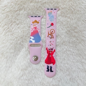 May include: A pink Apple Watch band with a Sleeping Beauty design. The band features images of Sleeping Beauty, her dress, a spinning wheel, a rose, and a wand. The band is made of silicone and has a silver buckle.