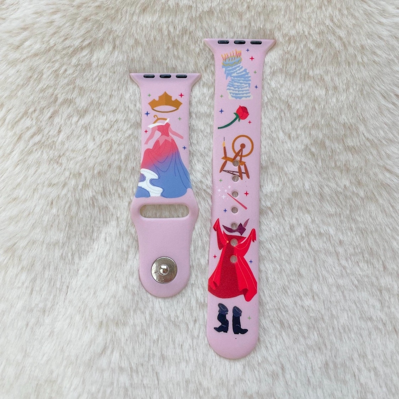 May include: A pink Apple Watch band with a Sleeping Beauty design. The band features images of Sleeping Beauty, her dress, a spinning wheel, a rose, and a wand. The band is made of silicone and has a silver buckle.