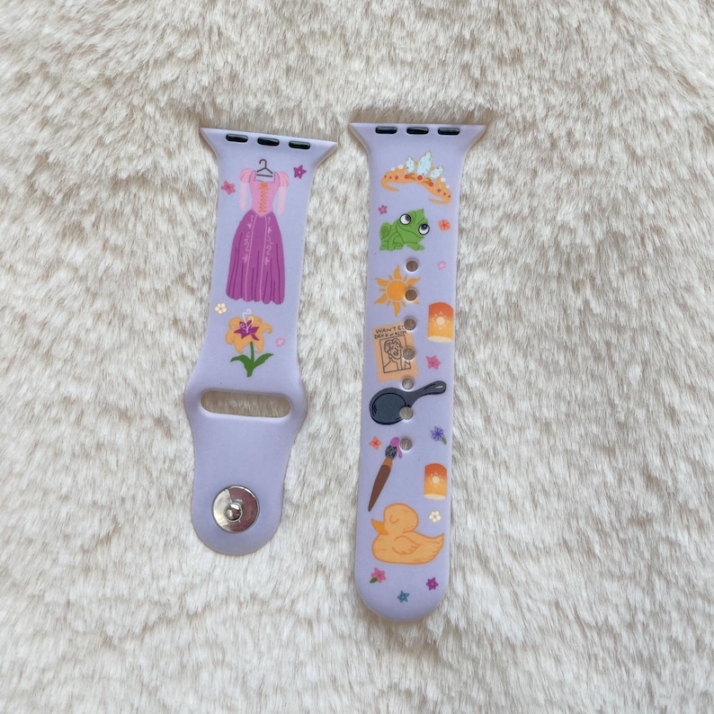 May include: Two lavender watch bands with colorful cartoon graphics. One band features a purple dress, flower, and stars. The other band has a frog, sun, lanterns, a frying pan, paint brush, duck, and stars.