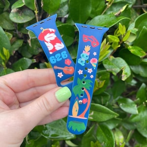 May include: Two blue Apple Watch bands with colorful designs. One band features a Hawaiian shirt, ukulele, and the number "626". The other band features a palm tree, surfboard, and a blue character with a flower crown.