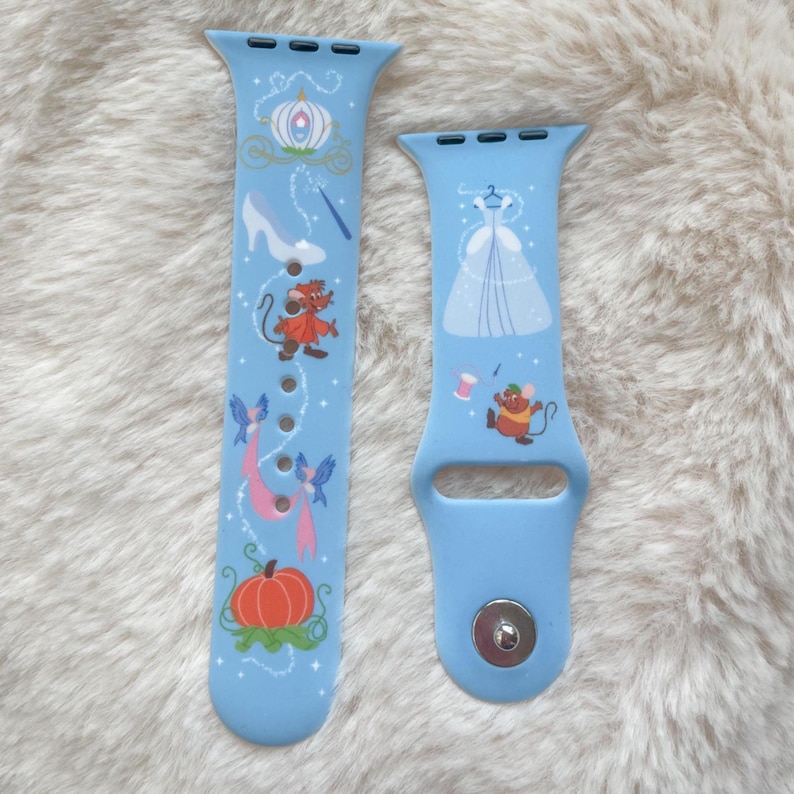 May include: Two light blue Apple Watch bands featuring Cinderella-themed illustrations. One band displays a pumpkin carriage, glass slipper, and fairy godmother's wand. The other band shows Cinderella's ball gown and a mouse with a spool of thread.