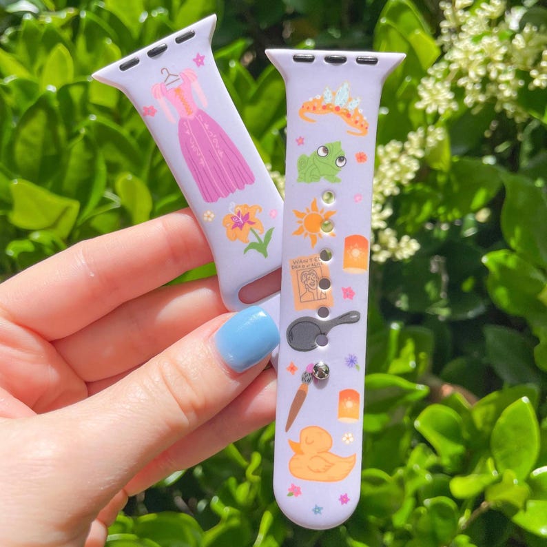 May include: Two lavender watch bands with colorful illustrations. One band features a pink dress, while the other showcases a frog, sun, and duck. The bands are decorated with stars and other themed elements.