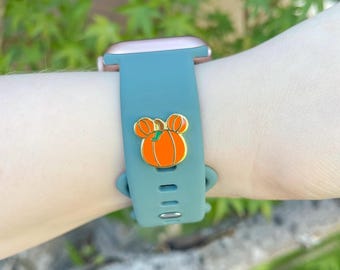 Könnte beinhalten: Türkisfarbenes Uhrenarmband mit goldenem Zifferblatt. Das Armband hat einen orangefarbenen Kürbis-Charm mit goldenen Konturen. Das Armband ist am Handgelenk befestigt.