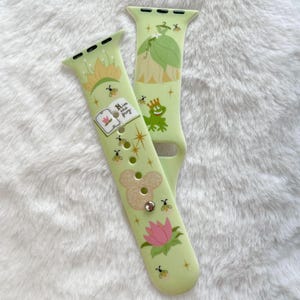 May include: A green and white Apple Watch band with a Disney Princess and the Frog theme. The band features a frog, a lily pad, a book with the text "Kiss the Frog", and a sparkly design.