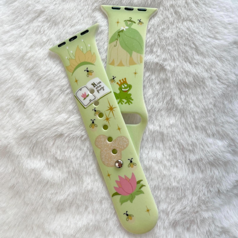 May include: A green and white Apple Watch band with a Disney Princess and the Frog theme. The band features a frog, a lily pad, a book with the text "Kiss the Frog", and a sparkly design.