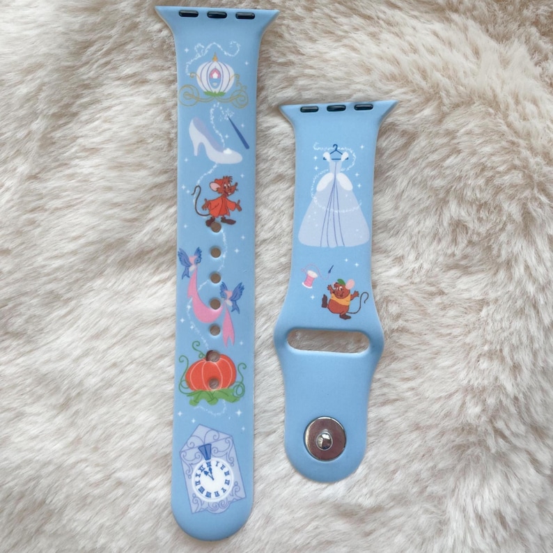 May include: Light blue watch bands featuring Cinderella-themed illustrations. One band displays a carriage, glass slipper, and clock. The other band shows a ball gown and a mouse. These bands are designed for a smartwatch.