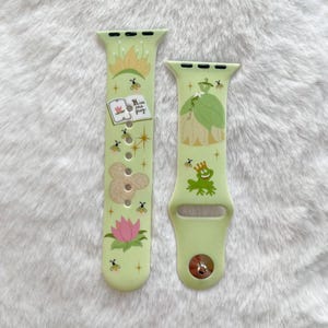May include: Two green Apple Watch bands with a princess and frog theme. The band on the left has a book with the text "His true love's frog" and a frog prince. The band on the right has a princess in a green dress.