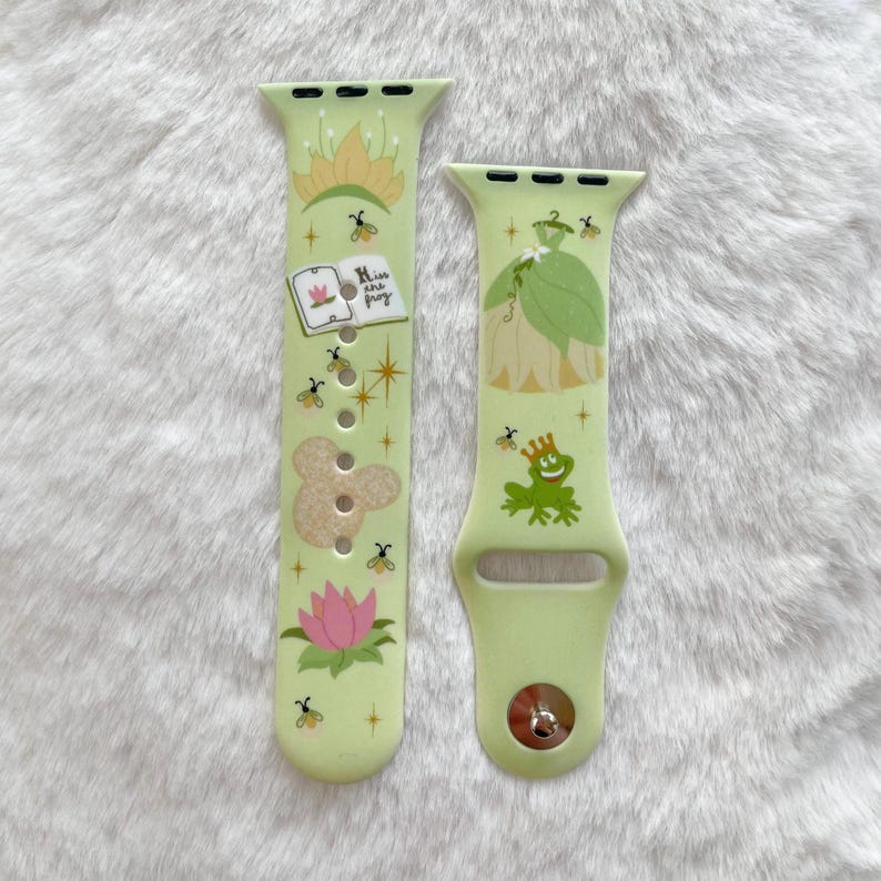 May include: Two green Apple Watch bands with a princess and frog theme. The band on the left has a book with the text "His true love's frog" and a frog prince. The band on the right has a princess in a green dress.