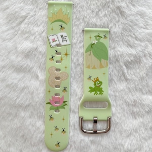 May include: Two green and white patterned watch bands with a princess and frog design. The band on the left has the text "Kiss the frog" on it. The band on the right has a silver buckle.